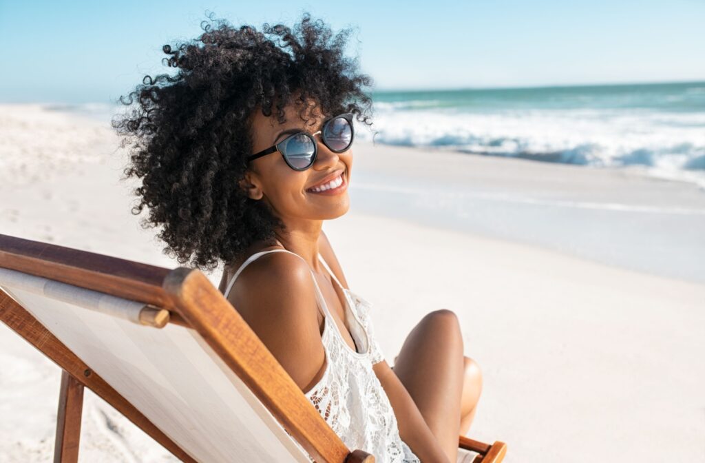 Young woman sitting at the beach in a lounger smiling & wearing polarized sunglasses.