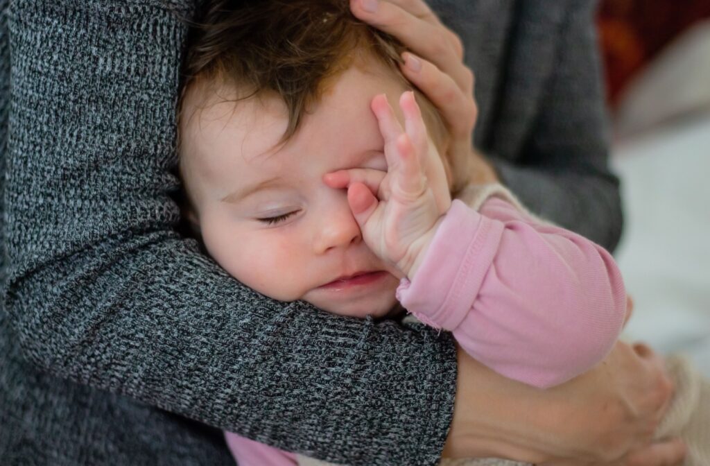 Tired baby in a pink romper rubbing her eyes. She is embraced in her mom's arms.