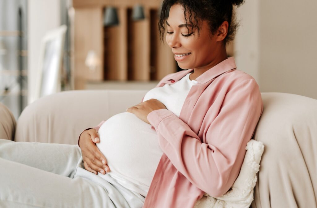 Smiling pregnant person relaxing on couch with hand resting on belly in a softly lit living space