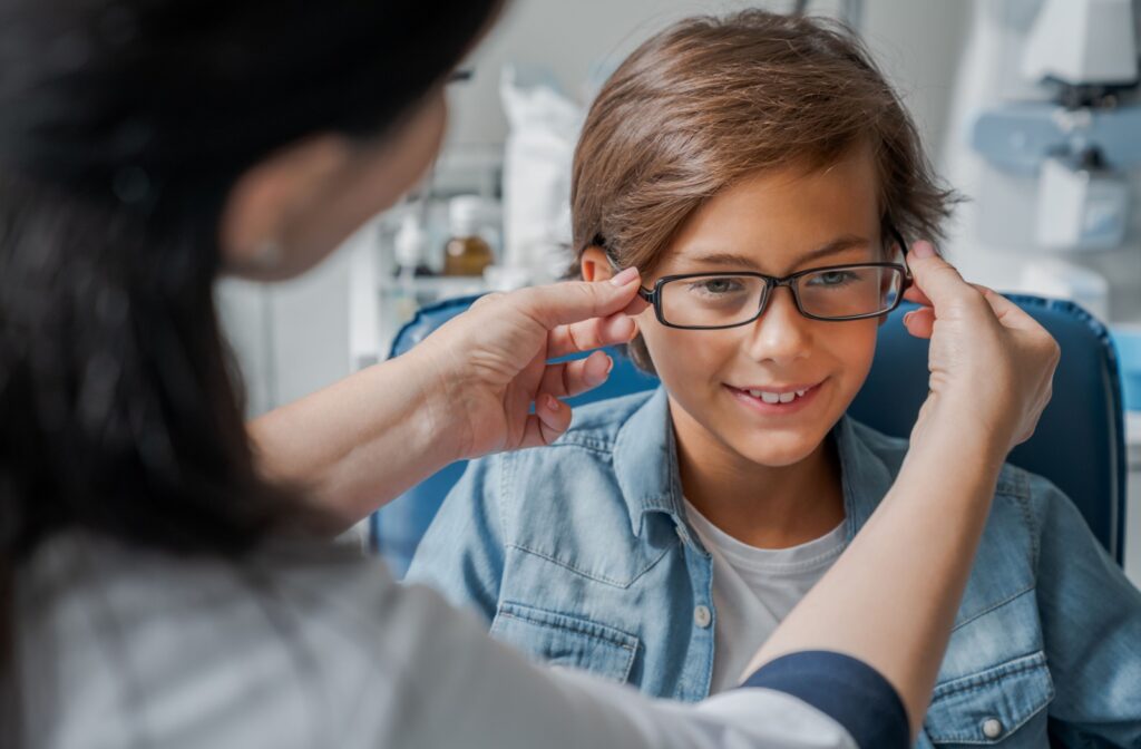 A child sits in an exam chair while an adult helps them try on a new pair of glasses.