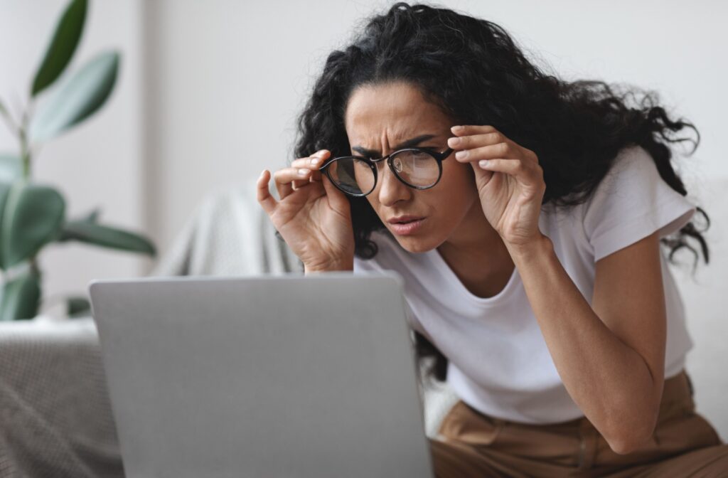 Woman squinting at a laptop screen while adjusting her glasses, having difficulty seeing clearly and focusing.